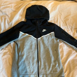 Nike ziphoodie i svart och grå - Nike hoodie med dragkedja i svart och grått. Jackan har huva, lång ärm och vit detalj över bröstet. Klassisk sportig look med Nike-logga på bröstet. Perfekt för dig som gillar streetwear och sportig stil.