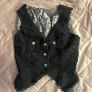 Svart mönstrad väst med dekorativa knappar & mönster - Säljer en svart väst med elegant mönster och silvriga dekorativa knappar framtill. Västen har två fickor med lock och ett snyggt paisleymönstrat innerfoder i grått och svart. Perfekt för att lyfta en outfit med en unik touch.