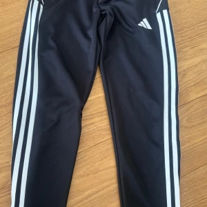 Svarta träningsbyxor med vita ränder från Adidas - Svarta träningsbyxor från Adidas med klassiska vita ränder längs sidorna och dragkedja vid benslut. Byxorna har en smal passform och är perfekta för sport eller vardag. Adidas-logga på ena benet.