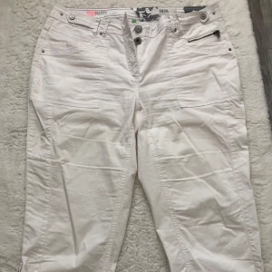 Vita byxor från Cecil med dragkedjedetalj - Säljer ett par vita byxor från Cecil med snygga detaljer som dragkedja vid fickan och flera knappar i midjan. Byxorna har raka ben och klassiska bakfickor. Perfekta för en fräsch och stilren look.