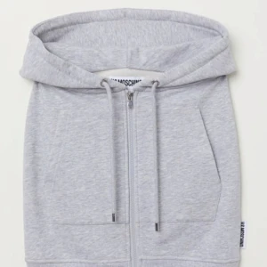 Grå kjol med dragkedja från Moschino x HM kollektionen - Säljer en grå kjol från Moschino i sweatshirtmaterial med dragkedja framtill, huva och snörning i midjan. Med tillhörande top. Kjolen har fickor på sidorna och en sportig look. Perfekt för dig som gillar streetwear och vill sticka ut.