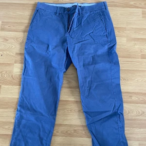 Blå chinos från Tommy Hilfiger - Snygga blå chinos från Tommy Hilfiger med klassisk passform. Byxorna har framfickor, bakficka med knapp och bälteshällor. Perfekta för en stilren och avslappnad look. Knappen i högerbackficka har gått sönder. Storlek 30/30