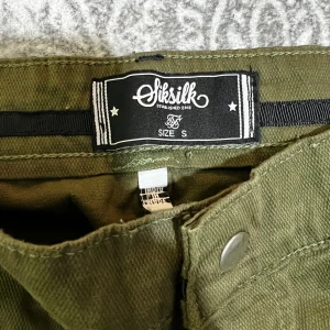 Olivgröna cargobyxor från SikSilk - Säljer ett par olivgröna cargobyxor från SikSilk med flera fickor på benen och dragkedjor vid fickorna upptill. Byxorna har en avslappnad passform och är perfekta för en streetwear-look.