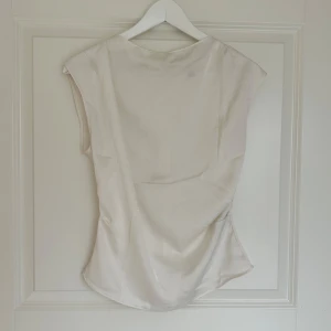 Vit satintopp - Säljer en elegant vit topp i satin med ärmlös design. Toppen har en mjuk glans och faller snyggt längs kroppen, perfekt för att lyfta vilken outfit som helst. Använt ett få tal gånger.