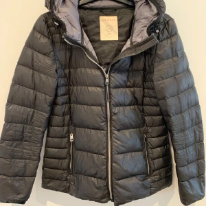 Svart pufferjacka från Esprit - Säljer en svart pufferjacka från Esprit med huva och dragkedja framtill. Jackan har två fickor med dragkedja och quiltad design. Perfekt för kyligare dagar och enkel att matcha med olika outfits. Finns ett hål i ena jackfickan.