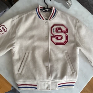 Beige varsityjacka med röd S och blå/röda detaljer - Snygg beige varsityjacka med stor röd S på framdidan och siffran 26 på ärmen. Jackan har blå och röda ränder på krage, ärmslut och midja. Stängs med knappar och har två fickor fram. Perfekt för en sportig och trendig look. Den är i xs men känns som s-m eftersom den är oversized. 