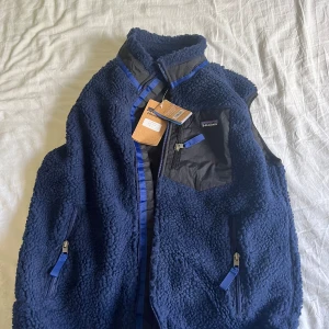 Blå fleeceväst från Patagonia - Mysig blå fleeceväst från Patagonia med svarta detaljer och dragkedjor. Västen har hög krage, bröstficka med dragkedja och två sidofickor. Perfekt för lager-på-lager och outdoor.
