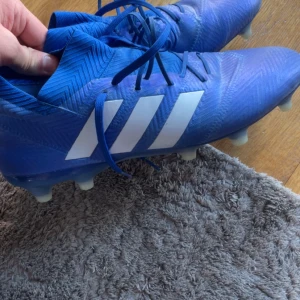 Blåa fotbollsskor från Adidas - Säljer ett par blåa fotbollsskor från Adidas med klassiska vita ränder på sidan. Skorna har snörning och är designade för att ge bra grepp på planen. Perfekta för dig som vill sticka ut på matchen! Använda 2 gånger. Skriv för fler bilder!