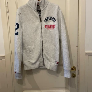 Grå ziptröja från Superdry - Säljer en ljusgrå ziptröja från Superdry med tryck i blått, vitt och rött framtill och på ryggen. Tröjan har hög krage, två fickor och muddar vid ärmslut och nederkant. Perfekt för en sportig look!