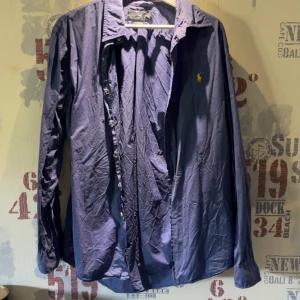 Blå skjorta från Polo Ralph Lauren - Säljer en klassisk blå skjorta från Polo Ralph Lauren med gul broderad logga på bröstet. Skjortan har lång ärm, knappar framtill och är i en custom fit-modell. Perfekt för dig som gillar stilrena plagg med ikonisk design.