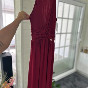 Vinröd långklänning med knyt i ryggen - Säljer en vinröd långklänning med smala axelband och snygg knytdetalj i ryggen. Klänningen har draperad midja och är ärmlös, vilket ger en elegant och luftig känsla. Perfekt för speciella tillfällen.