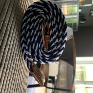 Blåvitt flätat bälte med bruna skinn-detaljer - Bra kvalitets bälte. Säljer ett snyggt blåvitt flätat bälte med bruna skinn-detaljer vid spännet och ändarna. Bältet har ett randigt mönster och passar perfekt till jeans. Justerbar längd tack vare det elastiska materialet.