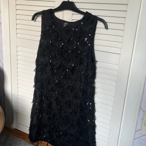 Svart paljettklänning - Säljer en svart ärmlös klänning från SHEIN med massor av svarta paljetter och fransdetaljer. Använd en enda gång och det finns inga tecken på användning! Klänningen har en rak passform och är täckt av glittrande paljetter som ger en festlig look. Perfekt för dig som vill sticka ut.