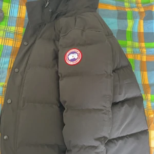 Svart dunjacka från Canada Goose med päls (Wyndham) - Säljer en svart dunjacka från Canada Goose med klassisk logga på ärmen och stor fluffig päls runt huvan. Jackan har knappar och dragkedja framtill och är perfekt för kalla dagar.