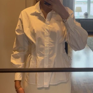 Vit skjorta från BikBok - Säljer en stilren vit skjorta från BikBok i storlek S. Skjortan har långa ärmar med resår vid ärmslut och markerad midja för en snygg siluett. Perfekt att styla med jeans eller kjol för en fräsch look. Använd en gång, som ny!!
