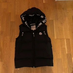 Svart dunväst med huva från Moncler - Säljer en svart dunväst från Moncler med huva och dragkedja framtill. Västen har två fickor med dragkedja och Moncler-logga på bröstet. Insidan av huvan har Moncler-tryck. Perfekt för lager-på-lager-stil.
