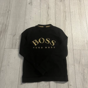 Svart sweatshirt från Hugo Boss - Svart sweatshirt från Hugo Boss med guldig BOSS-logga och texten HUGO BOSS på bröstet. Tröjan har rund halsringning och långa ärmar. Perfekt för en stilren och avslappnad look. Pris kan diskuteras!