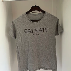Grå t-shirt från Balmain Paris - Säljer en stilren grå t-shirt från Balmain Paris med svart tryck framtill. T-shirten har rund halsringning och korta ärmar. Perfekt för en avslappnad och trendig look.