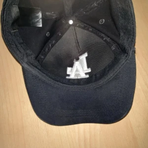 Svart LA keps med broderad logga - Säljer en svart keps med vit broderad LA-logga framtill och texten 'Los Angeles' baktill. Justerbart spänne bak för perfekt passform. Klassisk och stilren design, perfekt för dig som gillar sportiga accessoarer.