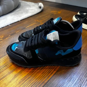 Valentino rock runners bara skor strl40 - Säljer ett par Valentino Rockrunner sneakers med svart och blått kamouflagemönster. Skorna har svarta snören, rund tå och platt sula. Materialmix av mocka och syntet med coola detaljer och ikonisk design.