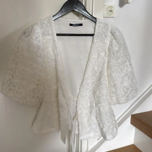 Vit spetsblus med puffärm från Gina Tricot - Säljer en vit blus från Gina Tricot med v-ringning, puffiga korta ärmar och omlottknäppning med knytband i midjan. Blusen är täckt av spets och har en romantisk känsla. Perfekt för dig som gillar detaljer och en feminin stil.