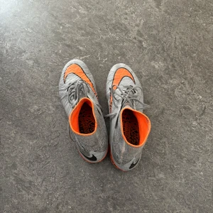 Grå och orangea Nike Hypervenom fotbollsskor - Säljer ett par gråa Nike Hypervenom fotbollsskor med orangea detaljer och högt skaft. Skorna har snörning och är designade för spel på gräs. Perfekta för dig som vill sticka ut på planen!