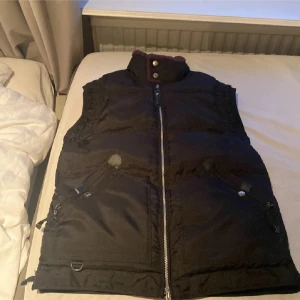 Svart dunväst med dragkedja - Säljer en svart dunväst med hög krage och dragkedja framtill. Västen har två fickor med knappar och detaljer i metall. Perfekt för lager-på-lager och håller dig varm under kyliga dagar.