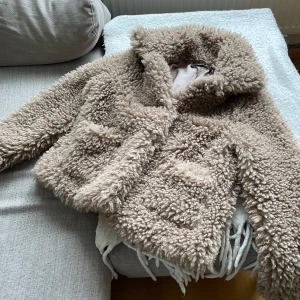 Beige teddyjacka från Zara Basic - Säljer en fluffig och trendig beige teddyjacka från Zara Basic. Jackan har stora fickor framtill, bred krage och är helfodrad. Perfekt för att hålla sig varm och snygg under kyliga dagar.