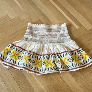 Vit kortkjol med mönster - Supersöt vit kortkjol med smockad midja och broderade gula och orange blommor längs fållen. Kjolen har volang och är gjord i ett lätt bomullstyg, perfekt för varma dagar. Färgglada detaljer och elastisk passform gör den både bekväm och trendig.