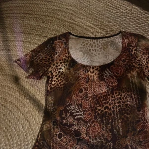 Brun mönstrad topp med leopard och blommor - Säljer en brun topp med korta ärmar och rund halsringning. Toppen har ett unikt mönster med leopardfläckar och blommor i olika nyanser av brunt och beige. Materialet är stretchigt och sitter tight mot kroppen.