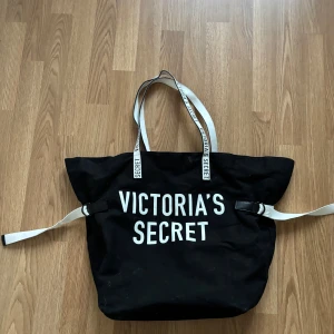 Svart shoppingväska från Victoria's Secret - Stor svart shoppingväska från Victoria's Secret med vita handtag och tydlig logga framtill. Väskan har innerficka och texten 'Love, Victoria' på insidan. Perfekt för att bära med sig mycket saker.