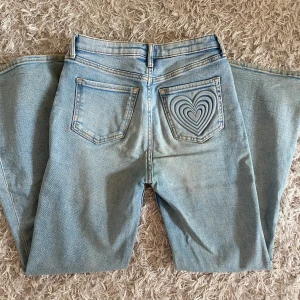 Ljusblå wide jeans med hjärtdetalj från H&M - Säljer ett par ljusblå jeans från H&M med breda ben och en unik hjärtdetalj på bakfickan. Jeansen har klassisk femficksdesign, normal midja och är tillverkade i denim. Perfekta för dig som vill sticka ut med en cool detalj!