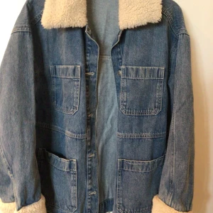 Jeansjacka med fuskpäls - Säljer en blå jeansjacka med fyra stora fickor framtill och mysig fuskpäls på krage och ärmslut. Jackan har en rak passform och stängs med knappar. Perfekt för kyligare dagar.