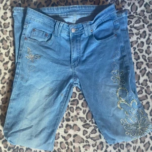 Flare jeans med detaljer - Säljer ett par blå jeans med broderade detaljer och rhinestones på framsidan. Byxorna har normal passform och vida ben. Lite skadade längst ner vid benen. Storlek S.