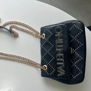 Svart quiltad axelväska från Valentino mario - Snygg svart axelväska från Valentino mario med quiltat mönster och nitar. Väskan har guldfärgad kedja och ett stort VALENTINO-tryck på locket. Perfekt för att lyfta din outfit med en lyxig touch.