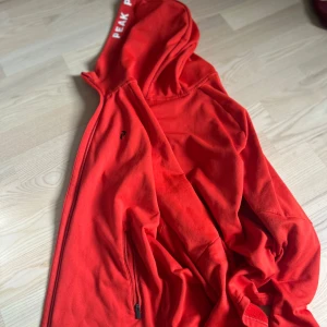 Röd hoodie med dragkedja från Peak Performance - Säljer en röd hoodie med dragkedja från Peak Performance. Jackan har huva med logga, långa ärmar och praktiska fickor med dragkedja. Perfekt för en sportig och avslappnad stil.