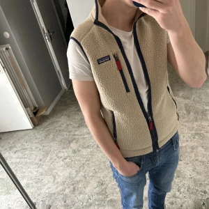 Beige fleeceväst från Patagonia - Snygg beige fleeceväst från Patagonia med marinblå detaljer och röda dragkedjor. Västen har hög krage, tre fickor med dragkedja. Mjuk och luftig som passar till många tillfällen. Storlek: small