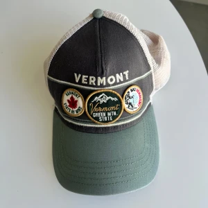 Vermont keps med broderade märken - Snygg keps med broderade märken och texten 'VERMONT' framtill. Framsidan är mörkgrå med grönt brätte och baksidan är beige mesh med justerbar knäppning. Märkena har motiv från Vermont och Green Mountain State.