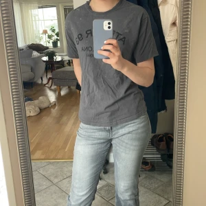 Oversized grå t-shirt med tryck  - Säljer en grå t-shirt från H&M Divided med svart texttryck 'ATELIER 93 BARCELONA Sants-Montjuic' på bröstet. T-shirten har rund hals och korta ärmar. Perfekt till en avslappnad stil.