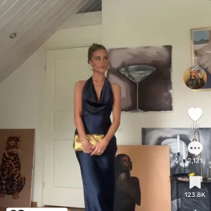 Mörkblå långklänning med halterneck - Säljer en elegant mörkblå långklänning med halterneck och djup v-ringning. Klänningen har en slät, glansig yta och en figurnära passform upptill som faller rakt ner. Perfekt för festliga tillfällen.