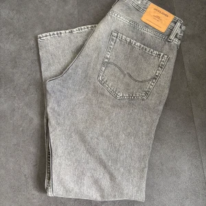 Grå jeans från Jack & Jones - Säljer ett par grå jeans från Jack & Jones i modellen Loose/Chris. Jeansen har klassisk femficksdesign, normal midja och lös passform. Perfekta för dig som gillar en avslappnad stil. Stängs med dragkedja och knapp.
