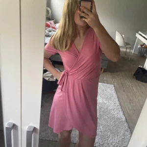 Rosa omlottklänning med knytband - Säljer en söt rosa omlottklänning med knytband i midjan. Klänningen har korta ärmar och v-ringad halsringning. Perfekt för varma dagar och enkel att styla med sneakers eller sandaler. Från vila. Strl xs 
