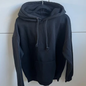 Svart hoodie med ficka - En klassisk svart hoodie med stor luva. Tröjan har en rymlig magficka och långa ärmar.