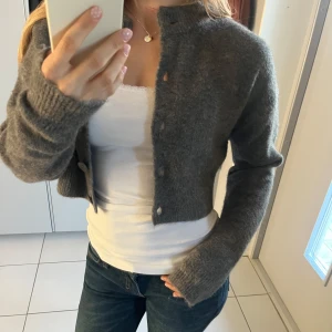 Grå croppad kofta med knappar - Mysig grå croppad kofta med långa ärmar och stora knappar framtill. Koftan är stickad och har en mjuk känsla.