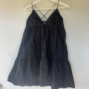 Svart klänning med smala axelband och volang - Svart klänning med smala axelband och v-ringning framtill. Klänningen har volangdetaljer och öppen rygg med snygg snörning. Perfekt för varma dagar och sommarens tillfällen.