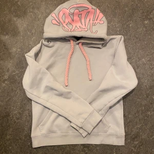 Synaworld OG Hoodie - Säljer denna grå och rosa hoodie från Synaworld, aldrig använd bara testad. Den har rep som snören och tryck på huvan. Mjuk insida och känguruficka framtill. Perfekt för en avslappnad stil. Har bara små hål vid ändan av kängrufickorna, men bara något man märker vid nära inspektion.