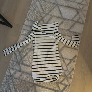 Randig ribbad klänning med offshoulder - Säljer en vit ribbad klänning med svarta horisontella ränder och offshoulder-design. Klänningen är tight och har långa ärmar. Perfekt för dig som gillar en stilren och trendig look.