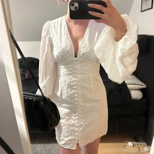 Vit kortklänning med puffärm från Adoore - Supersöt vit kortklänning från Adoore med v-ringning, puffiga långärmar och dekorativa knappar framtill. Klänningen har diskret prickigt mönster i tyget och markerad midja för en feminin siluett. Perfekt för sommarens alla tillfällen! En knappt har tyvärr lossnat längst ner men det är inget man lägger märke till 🥰🤩😇storlek 38 men passar också 36!