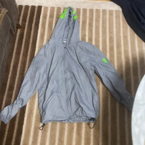 Grå vindjacka med huva från Risky - Grå vindjacka med huva och dragkedja från Risky Jackan har gröna detaljer på huvan och vänster ärm, samt snörning i nederkant. Perfekt för blåsiga dagar.
