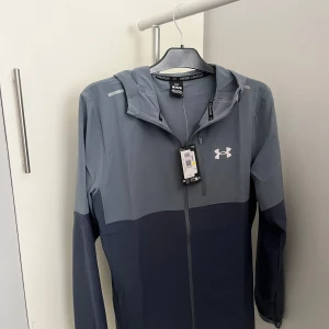 Under Armour jacka - Säljer en snygg tvåfärgad vindjacka från Under Armour i grått och mörkblått. Jackan är helt splitterny med pristagg och kvitto och aldrig använts, köpt från Jd.se. Med tanke på att den är slutsåld överallt och efterfrågan är så stor så är priset ganska rimligt men kan diskuteras.